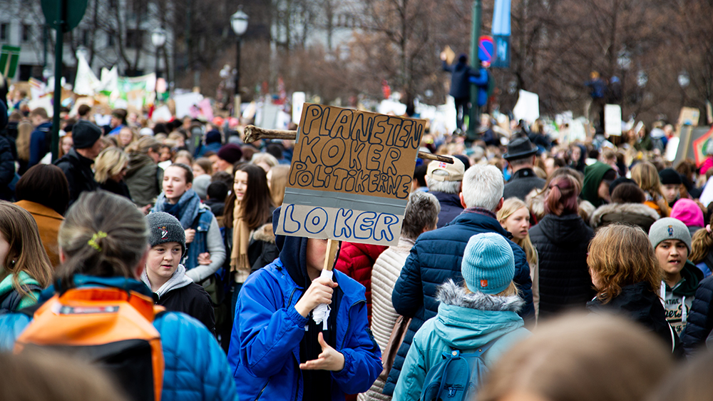 Barn og voksne demonstrerer. Gutt bærer plakat med påskrift: "Planeten koker. Politikerne loker"